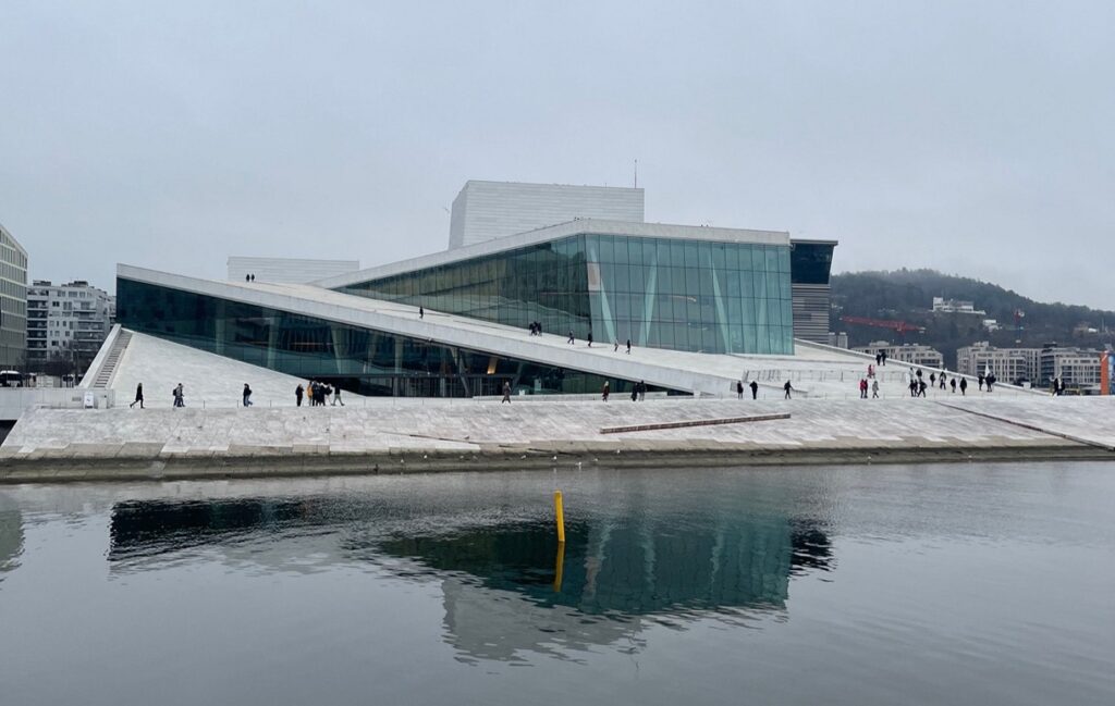 The image shows a modern building in Oslo, Norway. The sky is cloudy and the building is by the waters