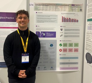 James Tawse stands in front of his academic poster. He is smiling and it is not possible to read the information on the poster