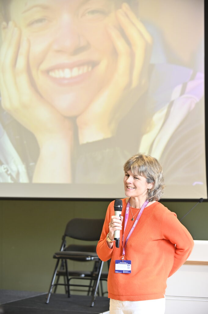A speaker stands on stage holding a microphone, with a large projected photograph visible on a screen behind them. The setting appears to be an indoor event or presentation space.
